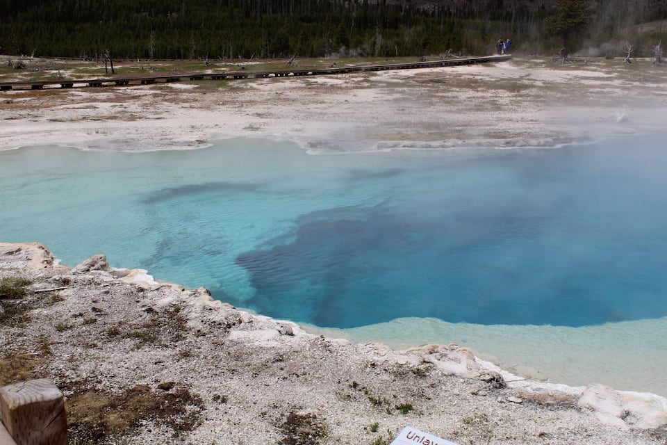 A geothermal area with clear blue water, a wooden walkway, and a forested background.