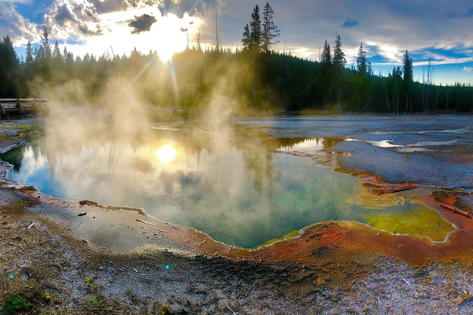 A geothermal area with a reflective pool, steam rising, and a forest in the background.