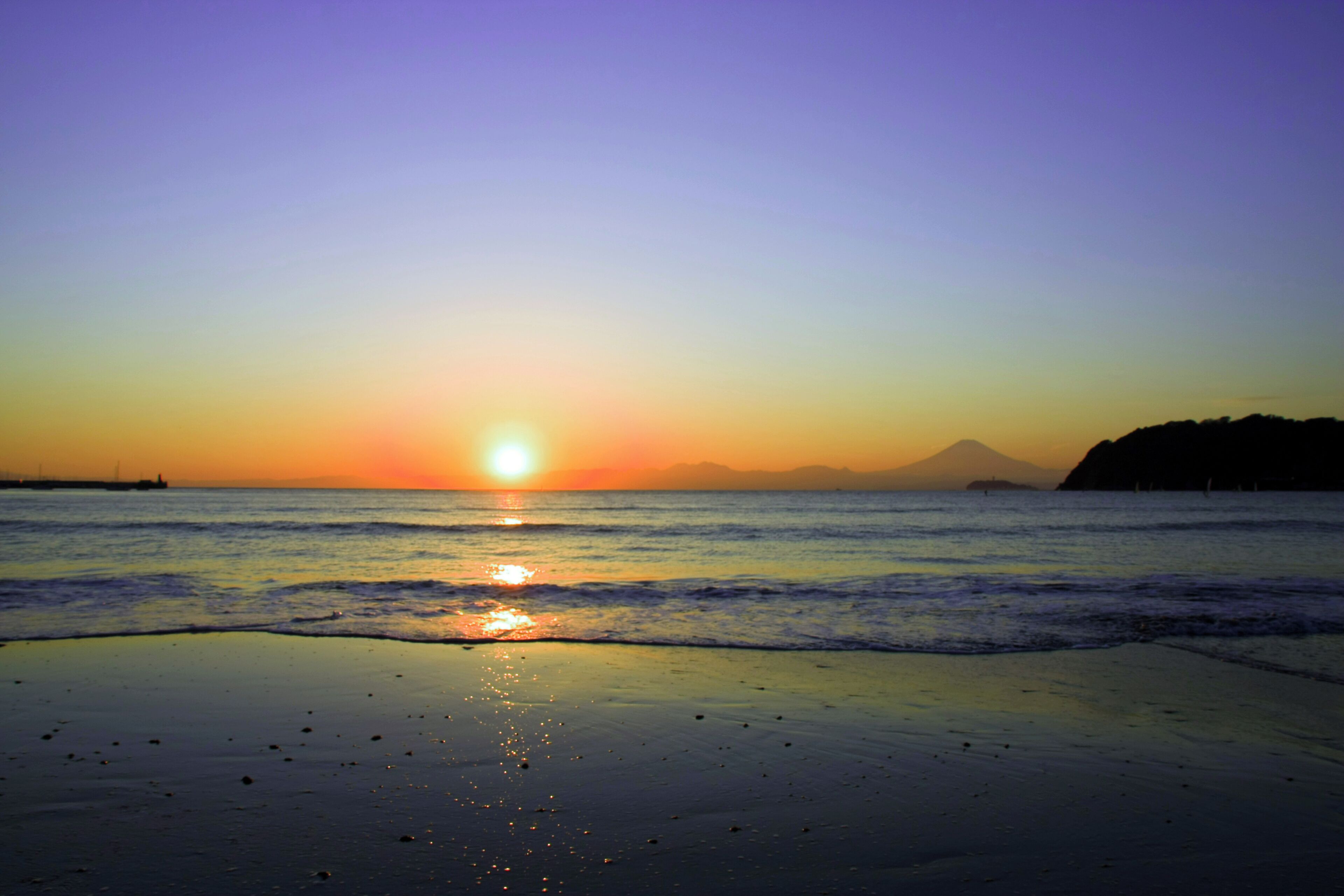Atardecer sobre una playa con olas y montañas a lo lejos.