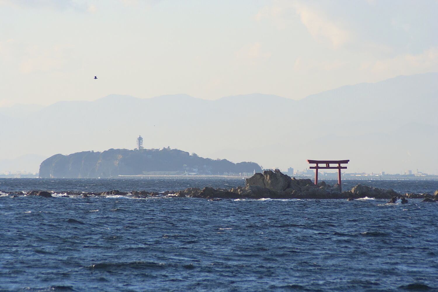 A coastal scene with a prominent red torii gate in the foreground, a rocky outcrop, and a distant island with a lighthouse.
