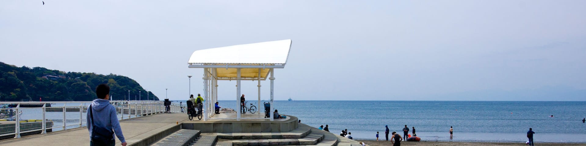 A coastal walkway leading to a beach with a white shelter and people walking and cycling.