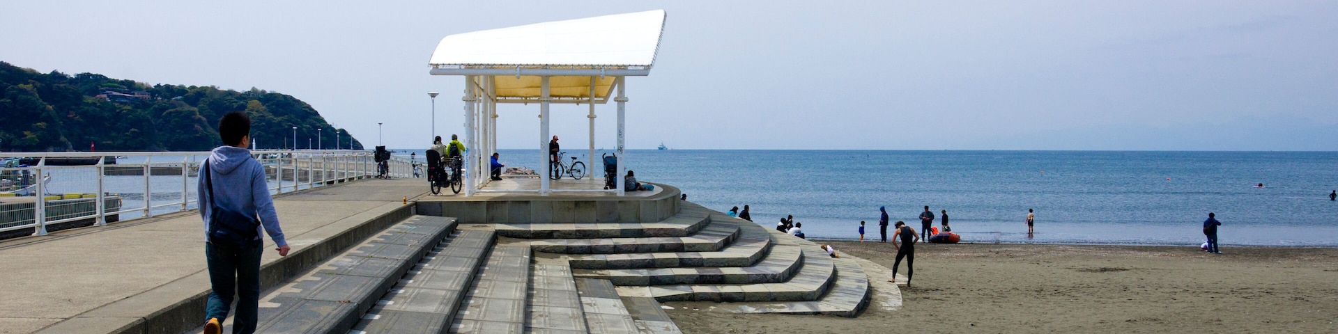 Un paseo marítimo que llega a una playa con un quiosco blanco y gente caminando y andando en bicicleta.