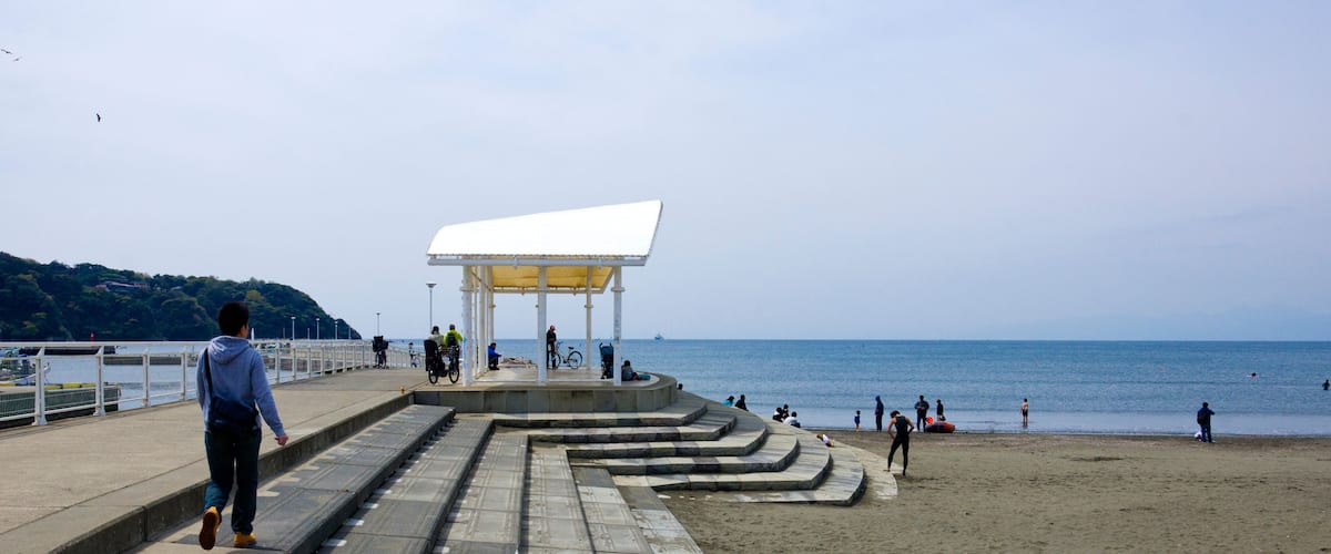 A coastal walkway leading to a beach with a white shelter and people walking and cycling.