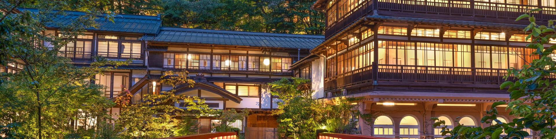 A traditional Japanese building with a wooden structure and multiple windows, surrounded by lush greenery and a red bridge leading to it.