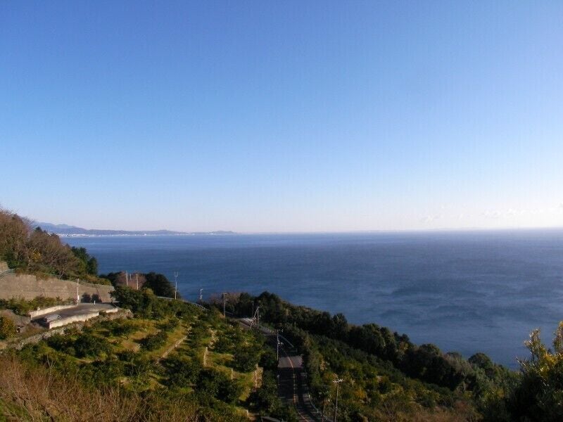 Paisagem costeira com vista clara do mar, relevo acidentado com vegetação e uma linha de costa distante com edifícios.