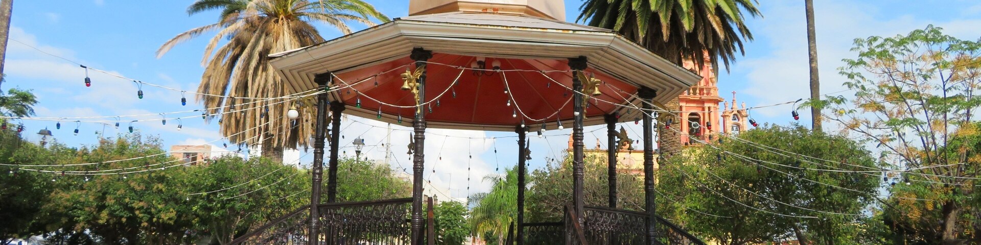 Un quiosco con techo abovedado y toldo rojo, rodeado de palmeras y una plaza con baldosas.