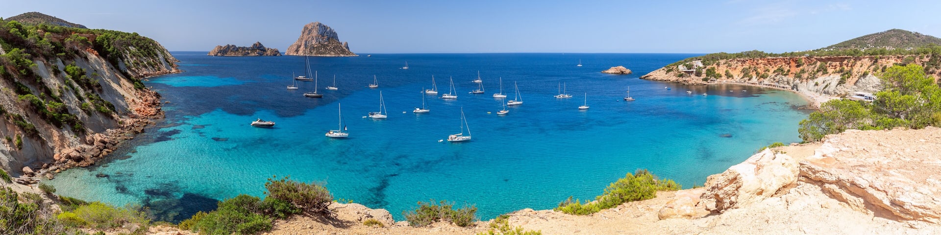 A coastal bay with clear turquoise waters, numerous sailboats, rocky cliffs, and a prominent rocky island in the distance.