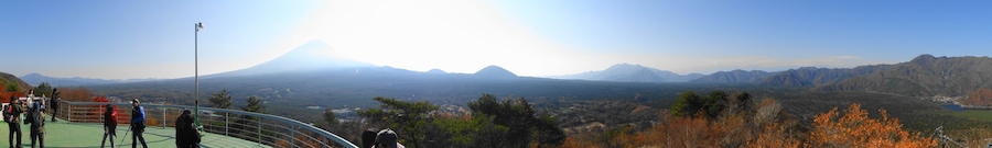 Paisagem montanhosa com um vulcão imponente ao longe, rodeada por árvores em tons de outono e um céu claro.
