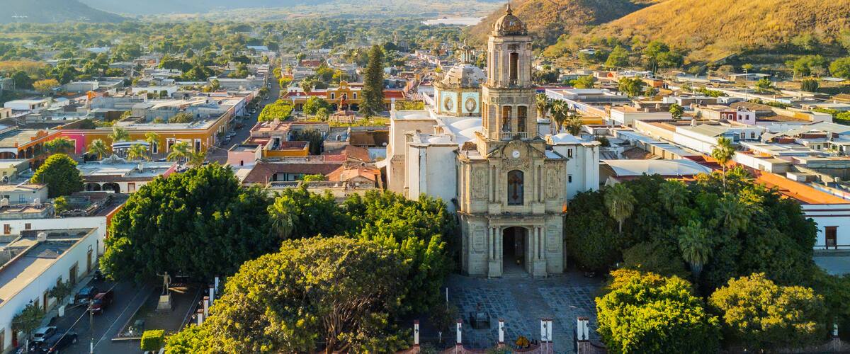 A town with a prominent church featuring a bell tower, surrounded by lush greenery and set against a backdrop of mountains.