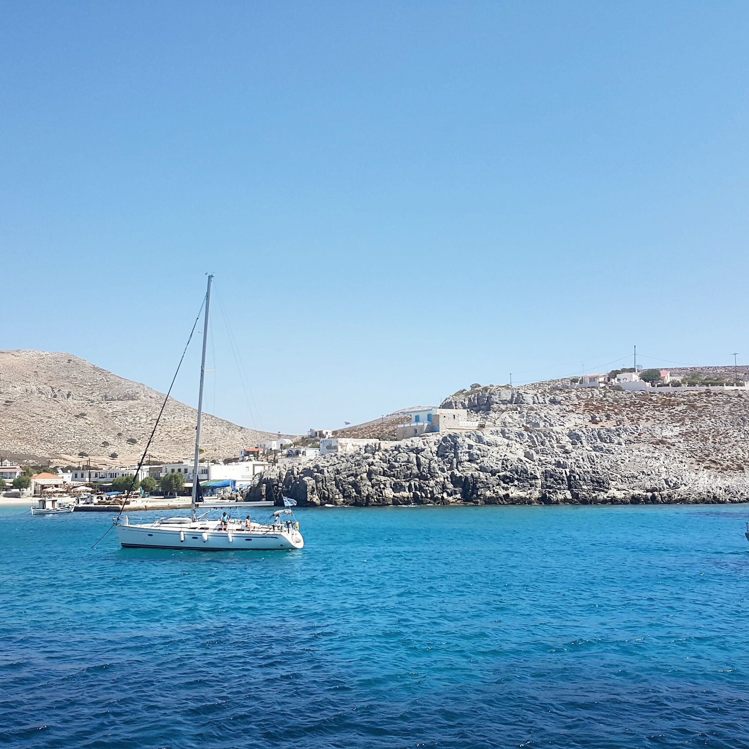 A sailboat on calm blue waters with a rocky coastline and buildings in the background.