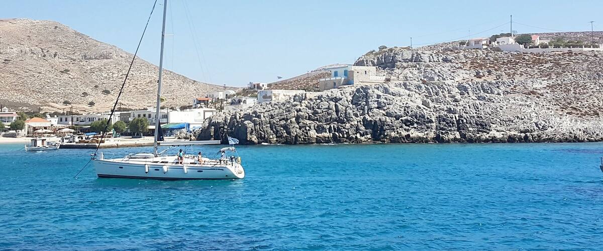 A sailboat on calm blue waters with a rocky coastline and buildings in the background.