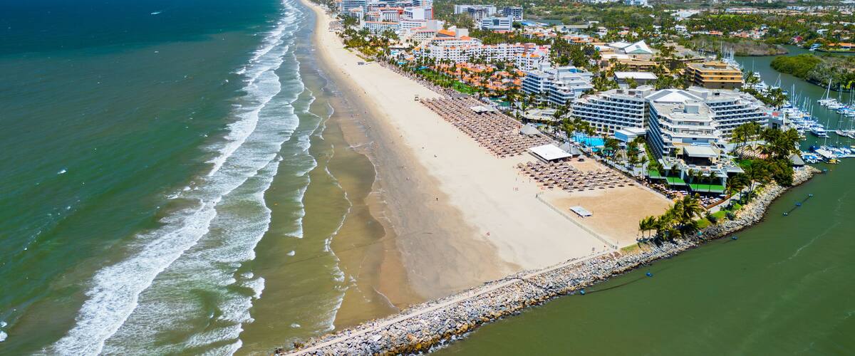 A coastal cityscape with a wide beach, a marina, and a backdrop of mountains.