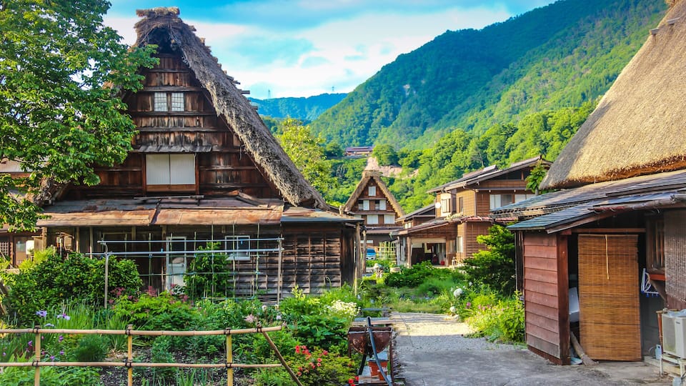 Traditional wooden houses with thatched roofs, a garden with various plants, and a mountainous backdrop.