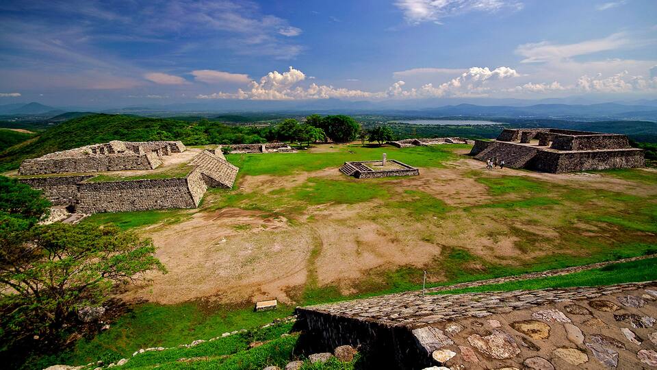 Ancient ruins with stepped pyramids and stone walls, surrounded by green hills and a clear sky.