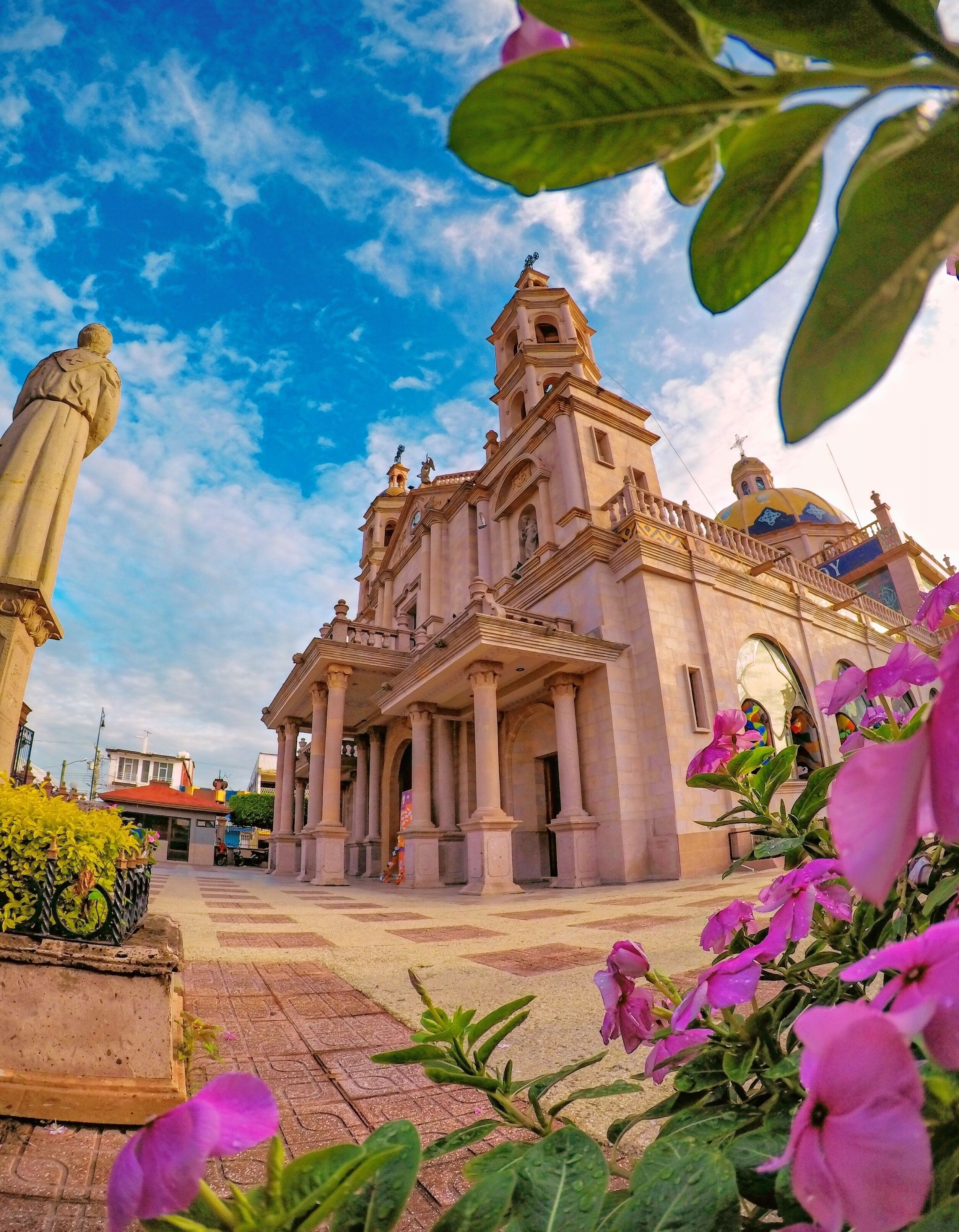 A historic church with a bell tower and dome, surrounded by a plaza with flowers and a statue.