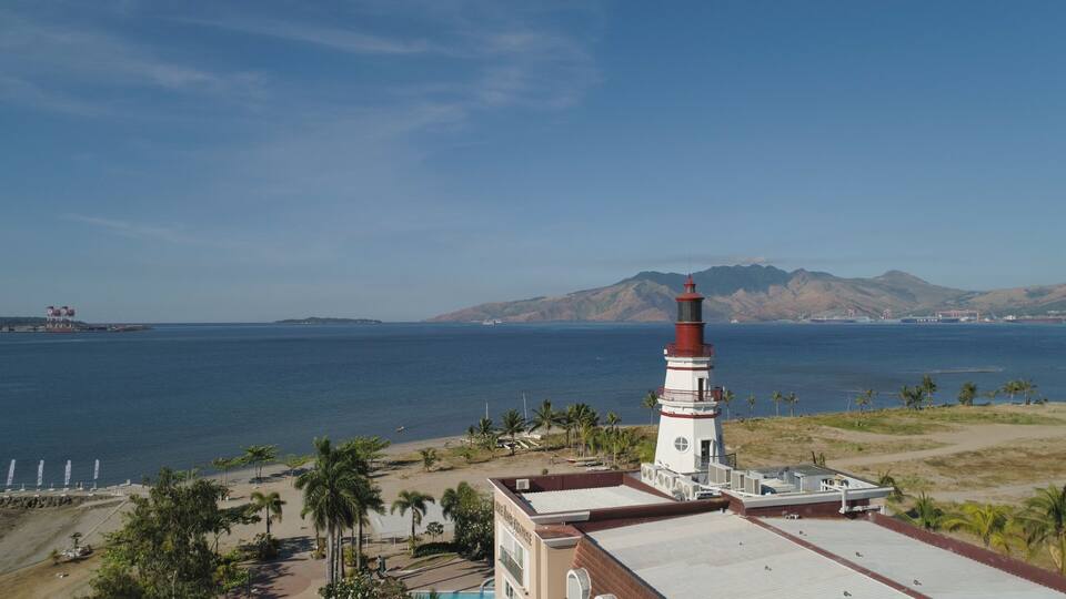 A lighthouse with a red top, a beach with palm trees, and mountains in the background.