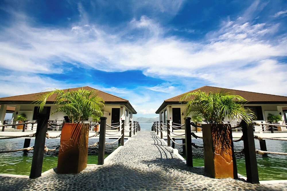 A pathway leading to two overwater bungalows with palm trees, surrounded by water and a clear sky.