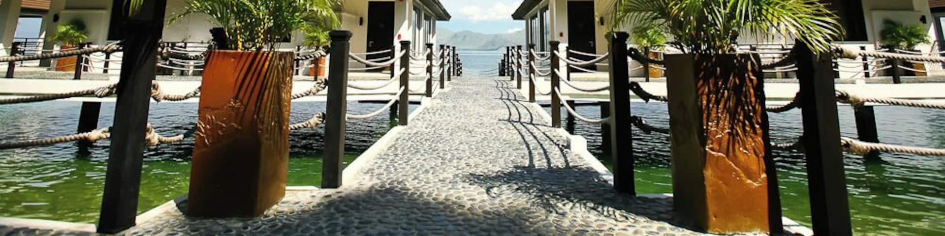 A pathway leading to two overwater bungalows with palm trees, surrounded by water and a clear sky.