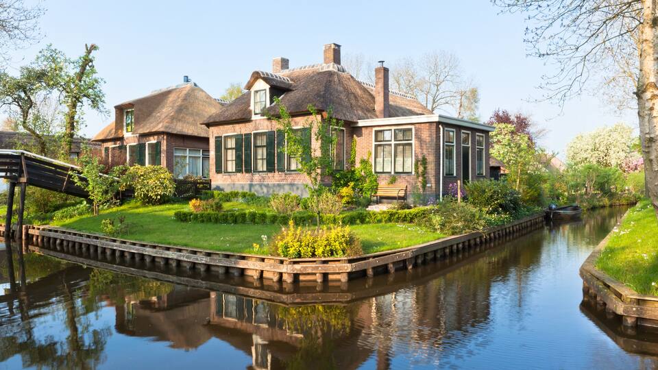A canal-side house with a thatched roof, surrounded by lush greenery and a calm waterway.
