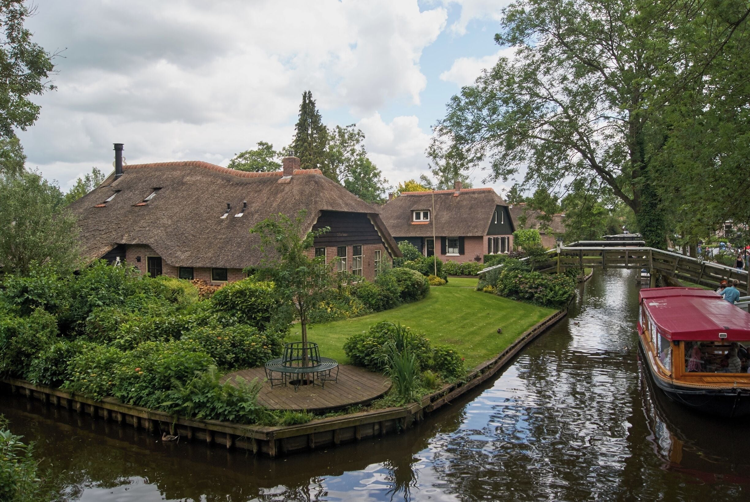 A canal with a wooden dock, a thatched-roof house, and a red boat.