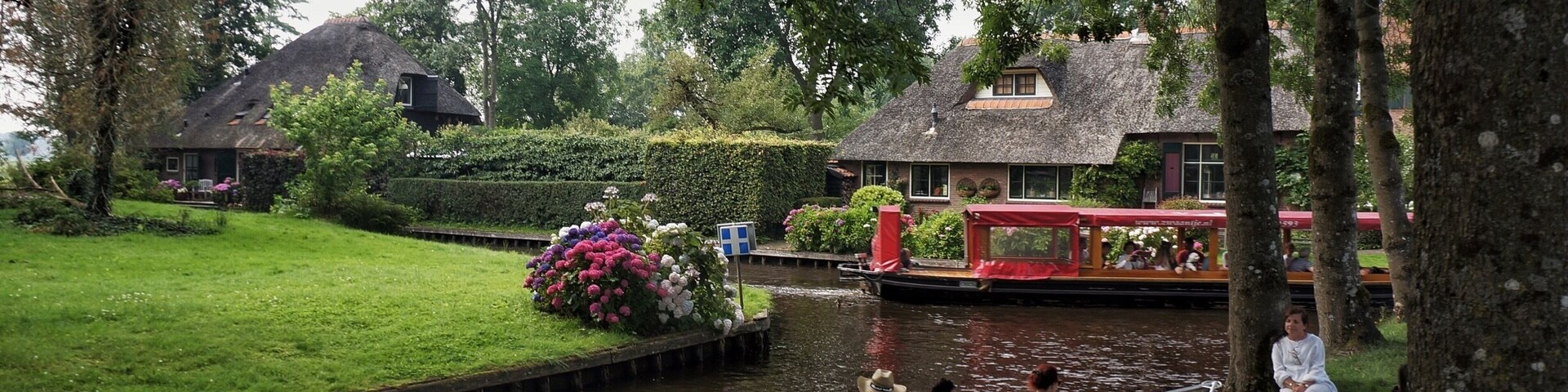Un canal con un bote, gente sentada en la orilla, y una casa roja al fondo.
