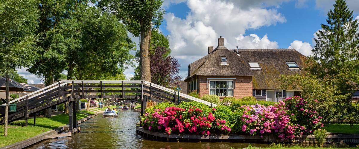 A canal with a wooden bridge, a house with a thatched roof, and blooming flowers along the water's edge.