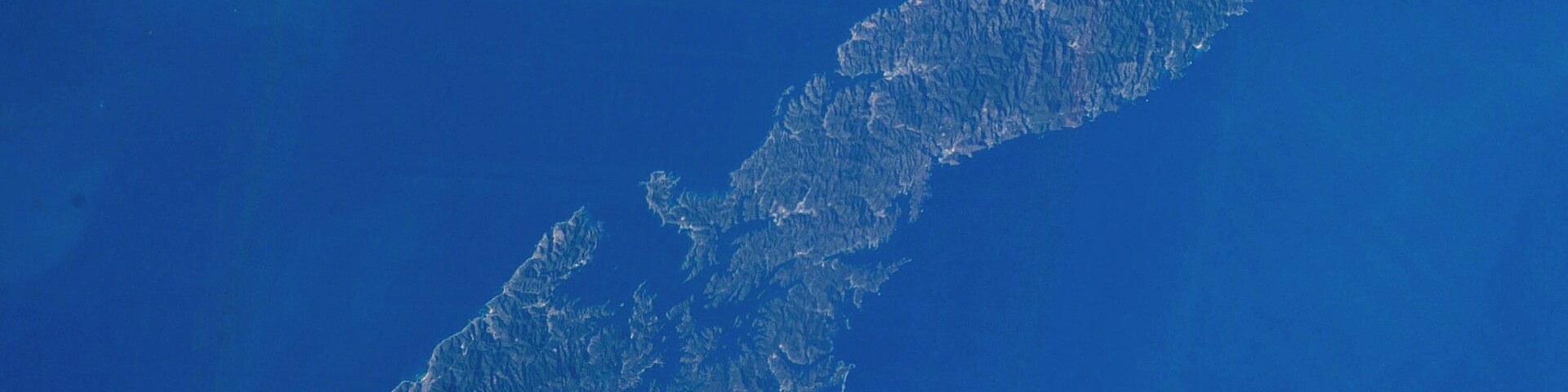 Vista de satélite de uma ilha extensa e alongada, com terreno acidentado e vegetação densa.