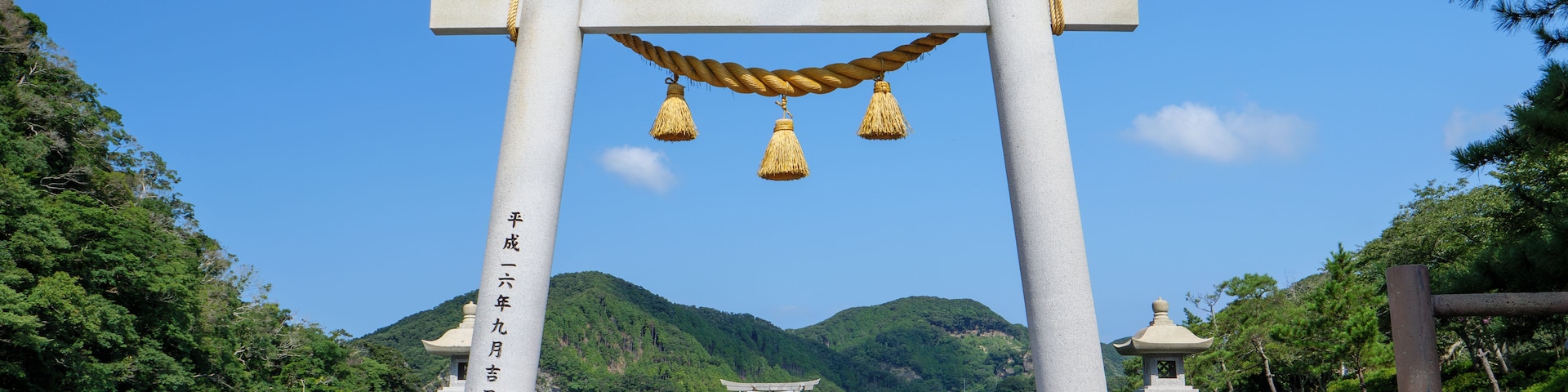 A traditional Japanese torii gate with a rope and tassels, a stone lantern, and a pagoda in the background, all set against a backdrop of a clear blue sky and lush greenery.