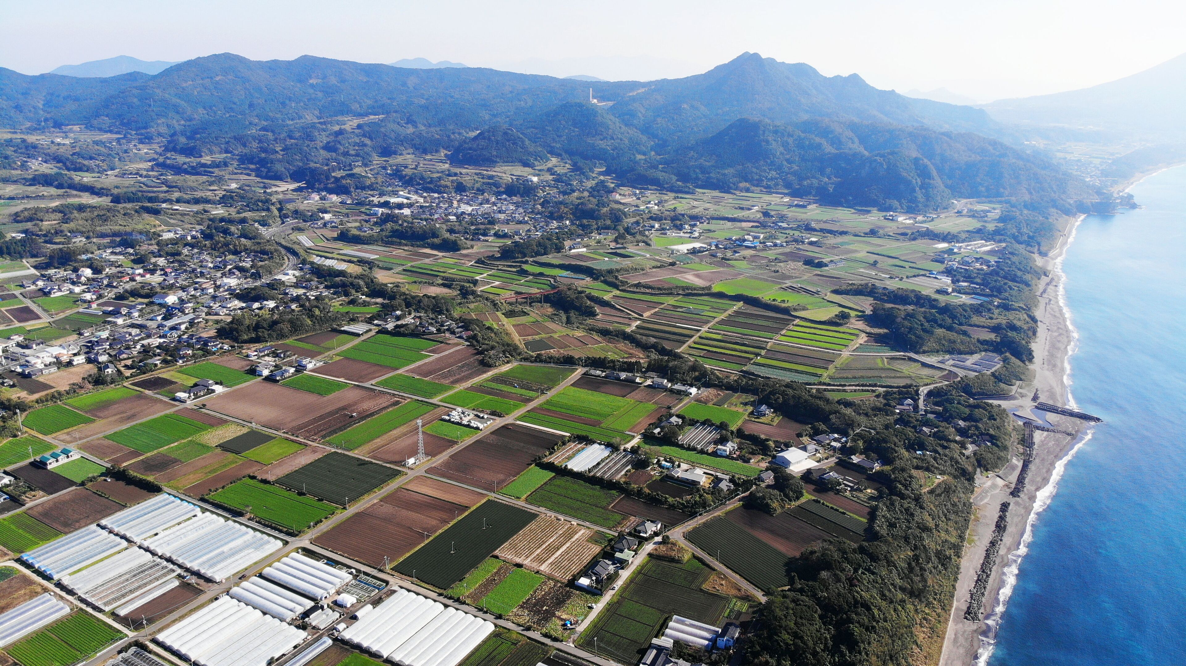An aerial view of a coastal town with agricultural fields, a beach, and mountains in the background.