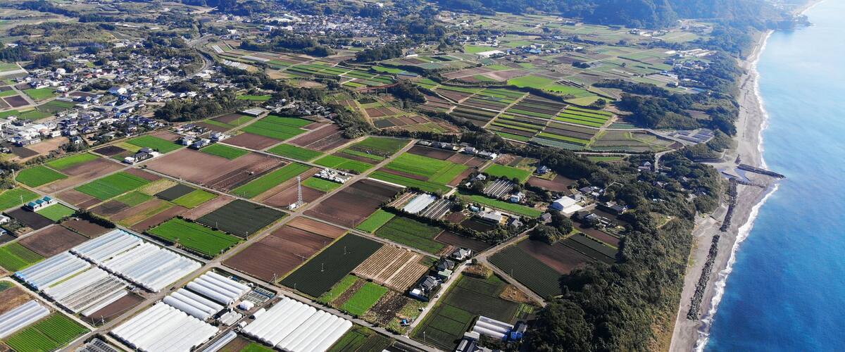 An aerial view of a coastal town with agricultural fields, a beach, and mountains in the background.