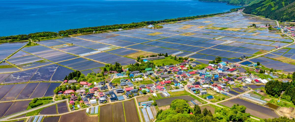 An aerial view of a coastal village with solar panel fields and a mountain range in the background.
