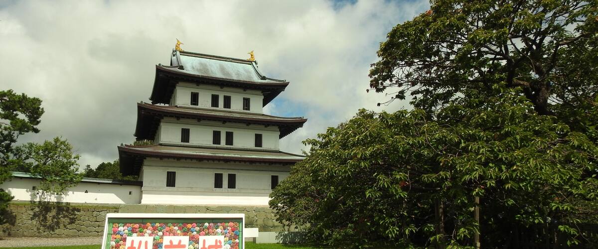 A traditional Japanese building with a multi-tiered roof and golden finials, surrounded by lush greenery and a stone wall.