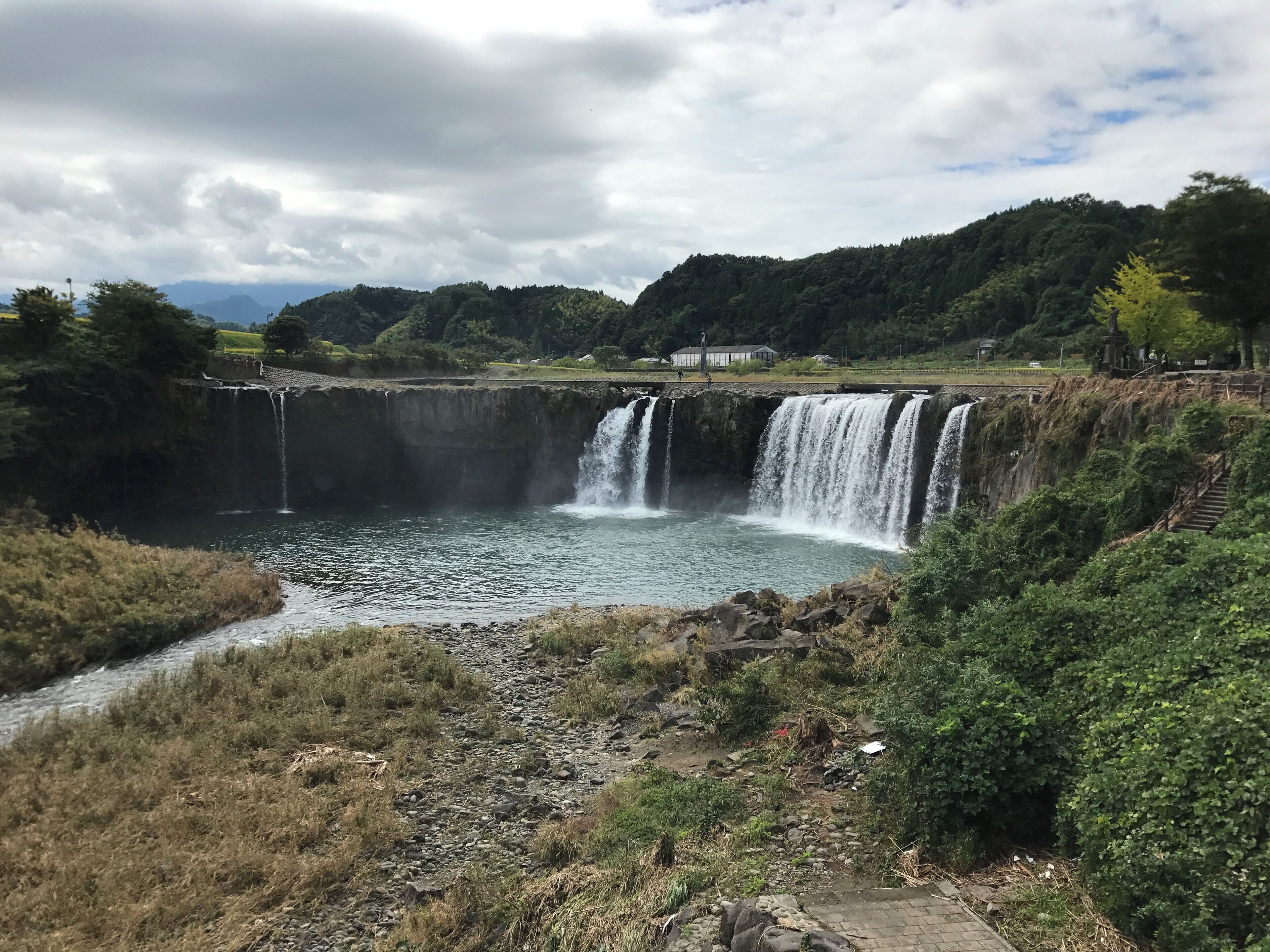 A waterfall cascading into a river, surrounded by greenery and a cloudy sky.