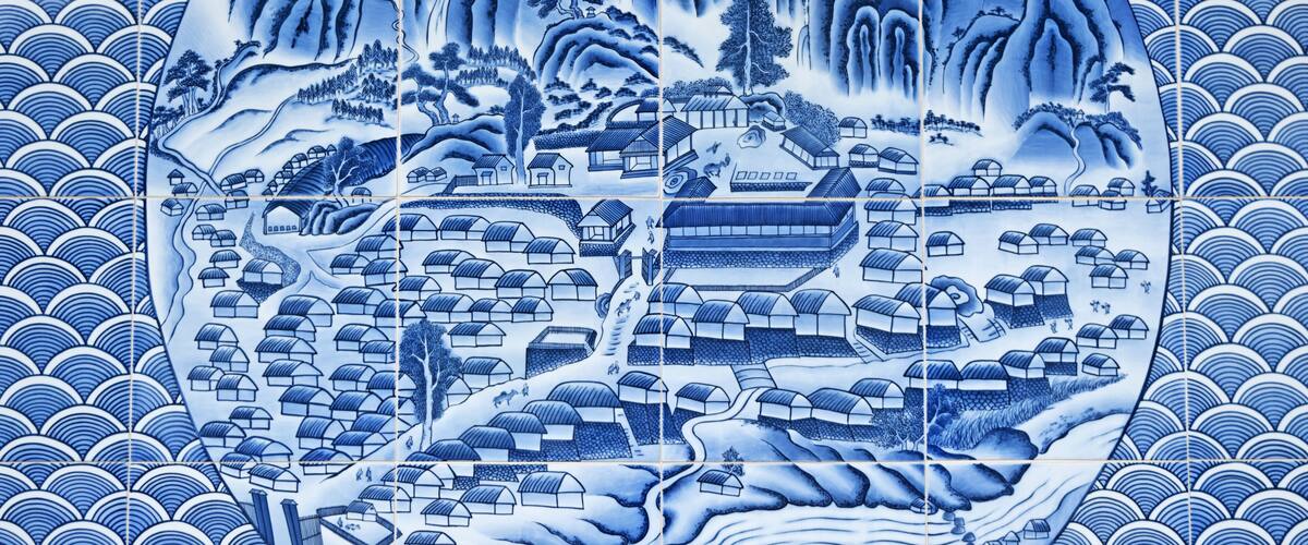 A blue and white ceramic tile with a traditional Japanese landscape scene, including mountains, trees, and houses.