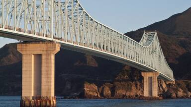 Un pont en acier à structure en treillis qui enjambe une étendue d’eau, soutenu par de grands piliers en béton, avec une chaîne de montagnes en arrière-plan.
