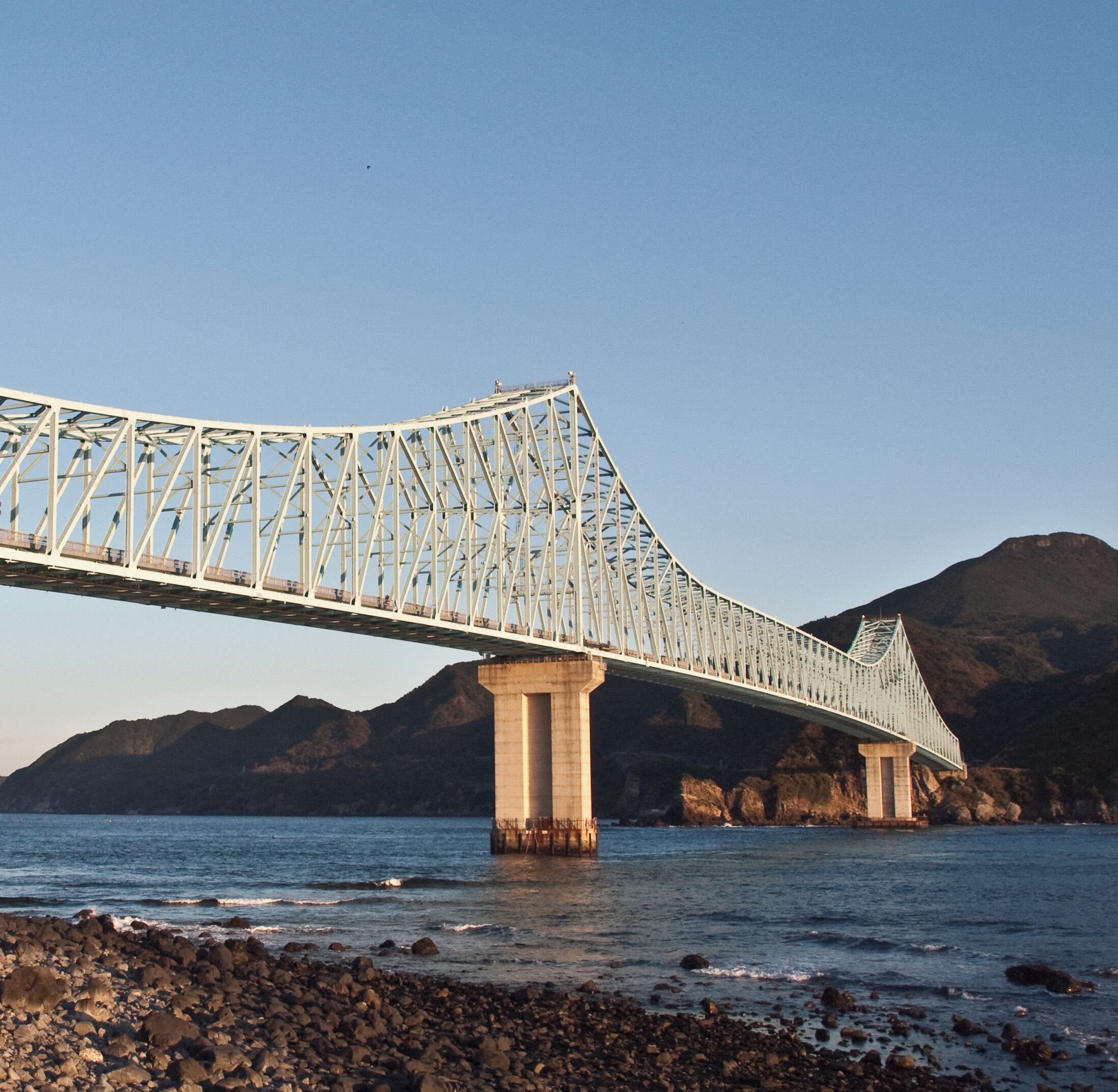 A large suspension bridge spanning a body of water with rocky shores and mountains in the background.