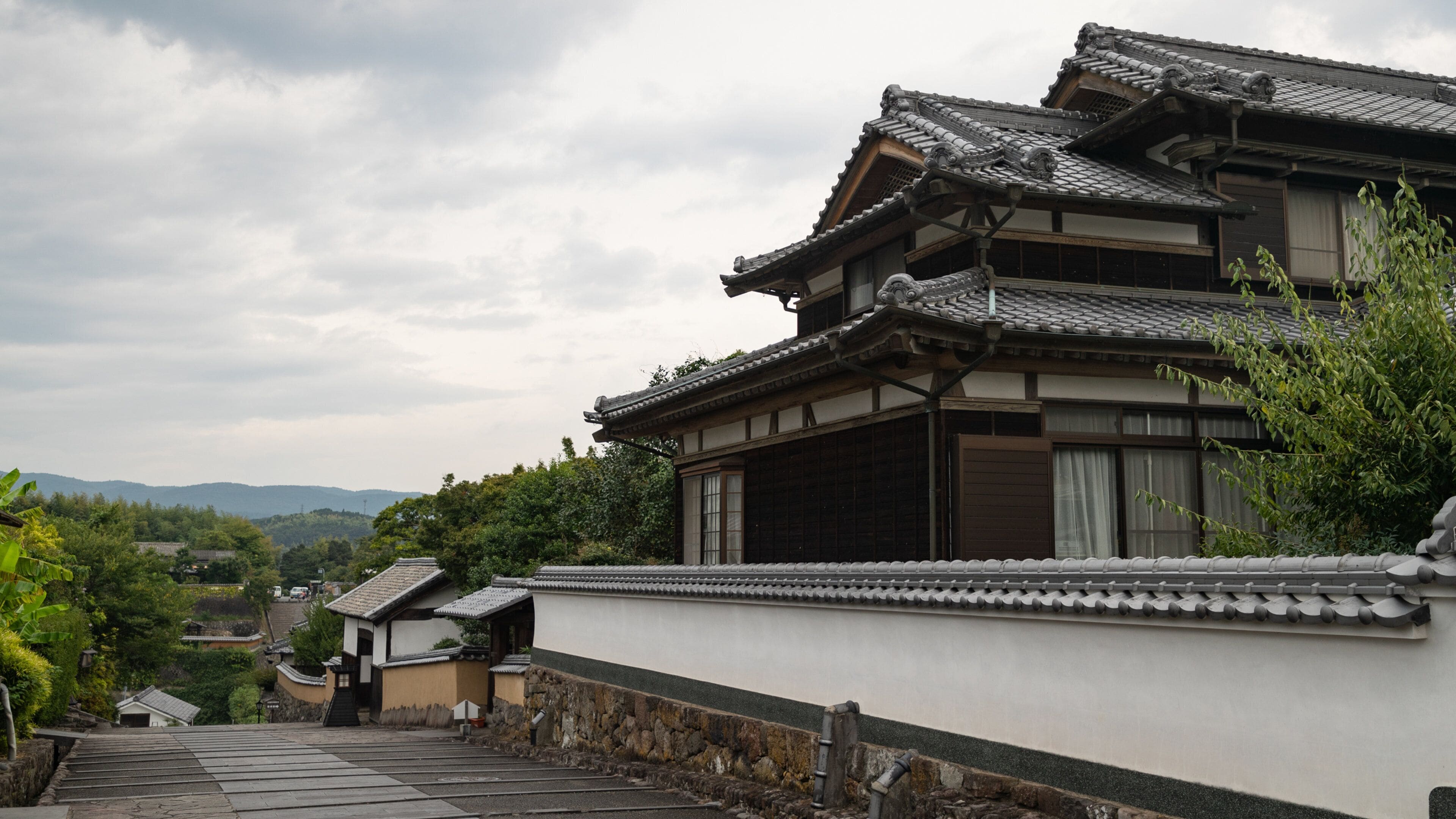 A traditional Japanese building with a tiled roof and wooden structure, surrounded by greenery and a stone pathway.