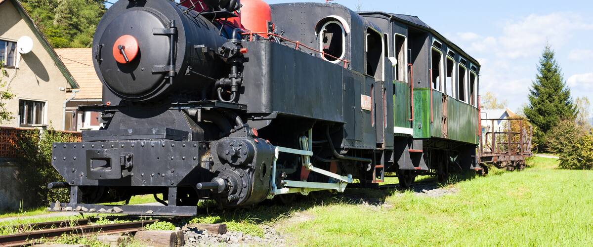 A vintage steam locomotive with a red front, green passenger carriages, and a caboose, on a rural railway track.