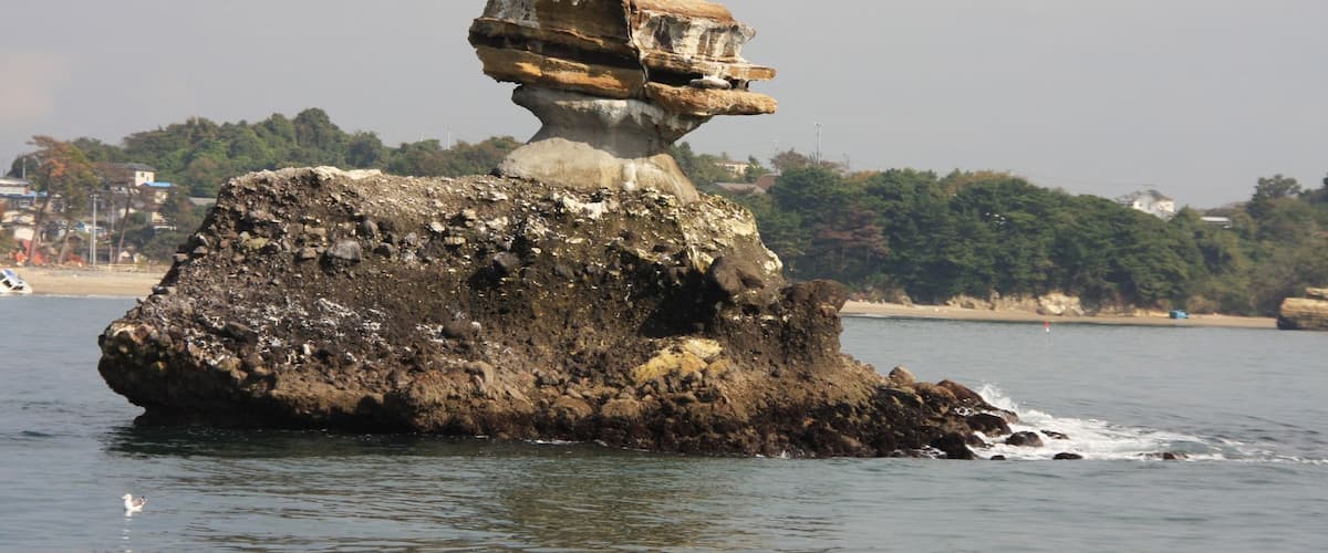 A rock formation with a balanced rock on top, surrounded by water and a distant shoreline with buildings.