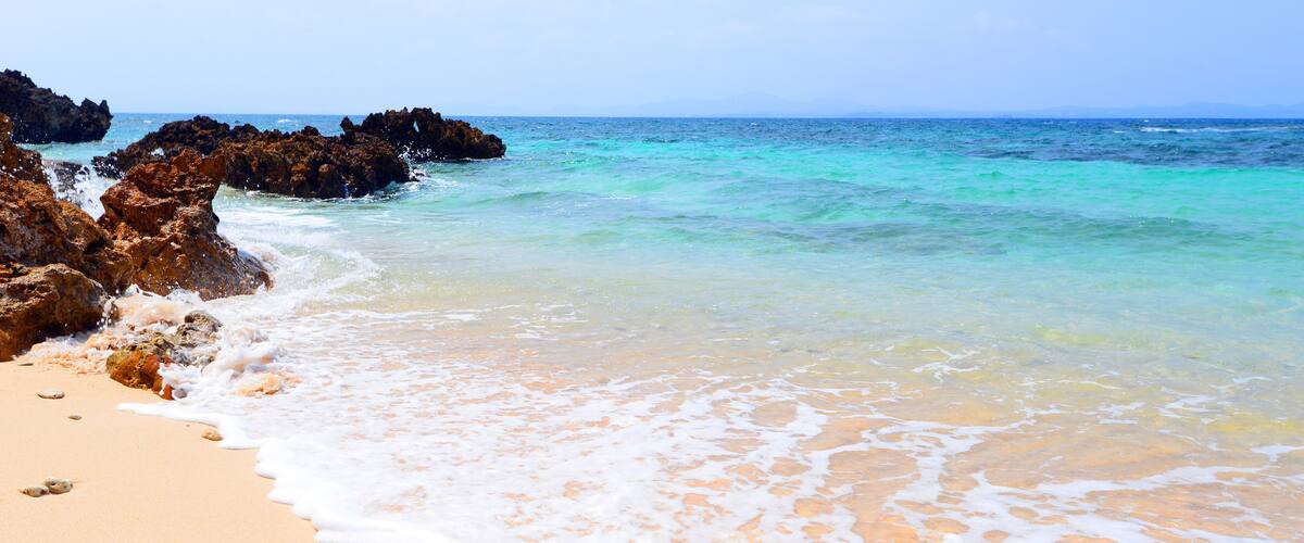 A sandy beach with gentle waves, clear turquoise water, and rocky outcrops.