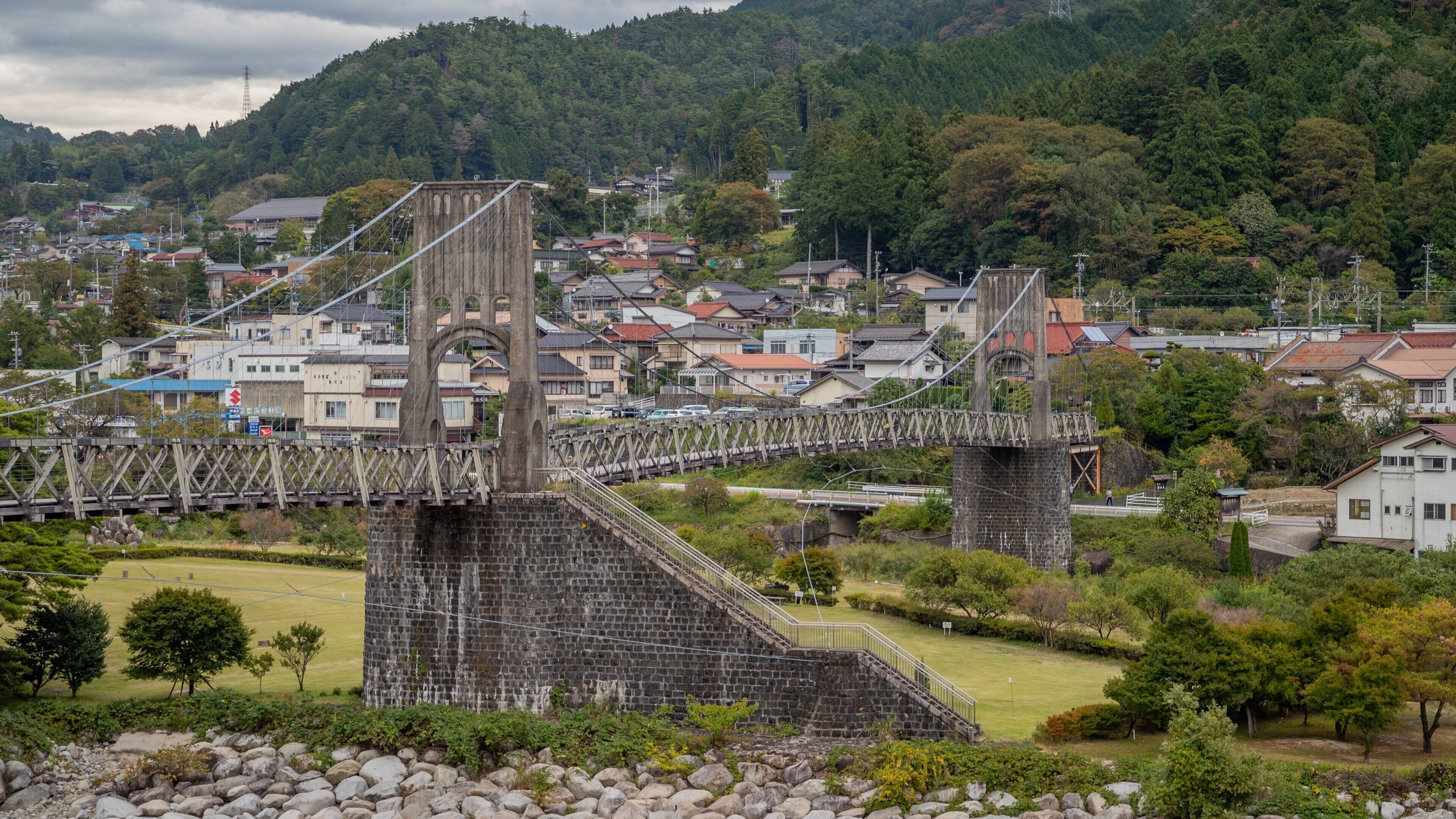 A stone bridge with a wooden walkway over a river, surrounded by greenery and residential houses.