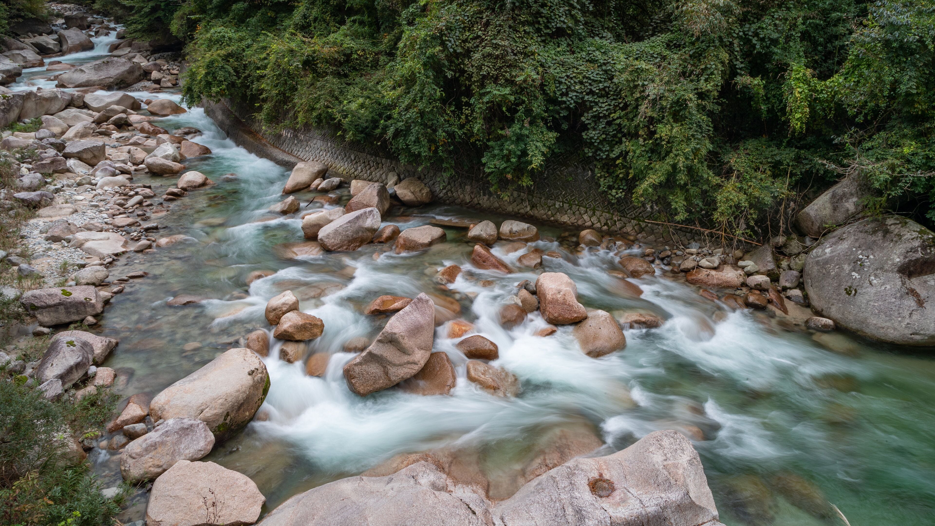 A river with clear water flowing over rocks, surrounded by lush greenery and a stone wall.