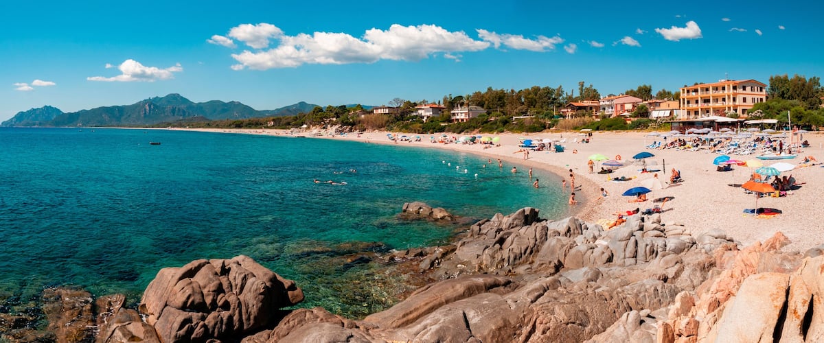 A coastal beach with clear turquoise waters, rocky shoreline, and buildings in the background.