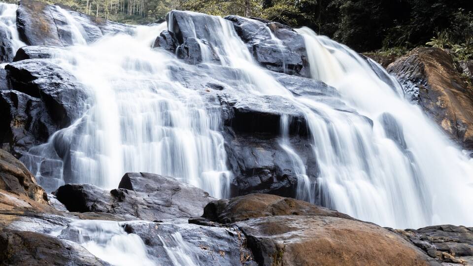 A multi-tiered waterfall cascading over large rocks in a forested area.