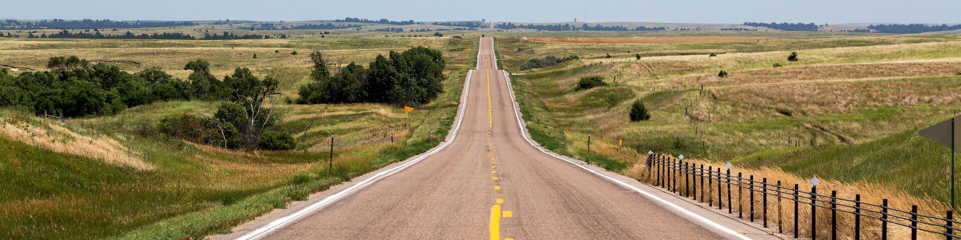 A straight road with a yellow center line, flanked by grassy fields and a wooden fence on one side.