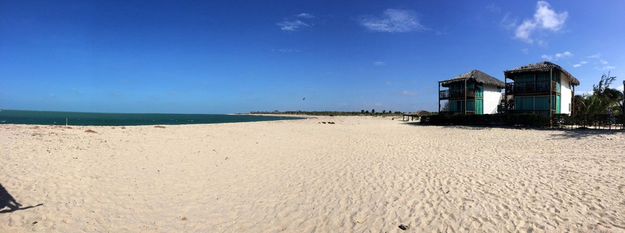 Uma praia arenosa com um edifício de telhado de palha e céu azul límpido.