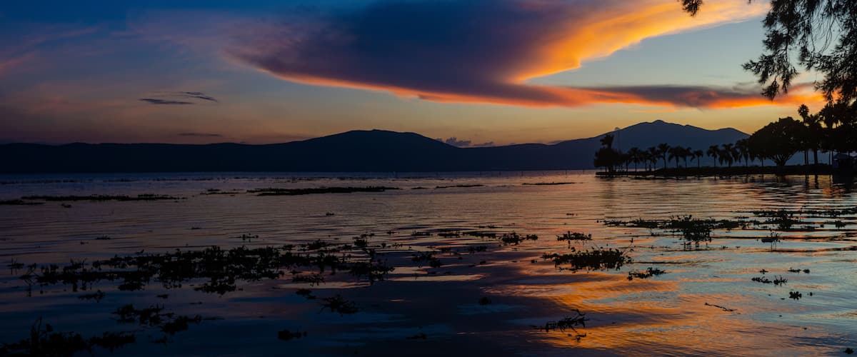 A sunset over a calm lake with distant mountains and palm trees.