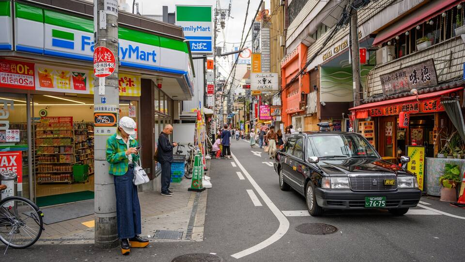 Uma rua movimentada com diversos estabelecimentos, incluindo uma loja FamilyMart e um restaurante.