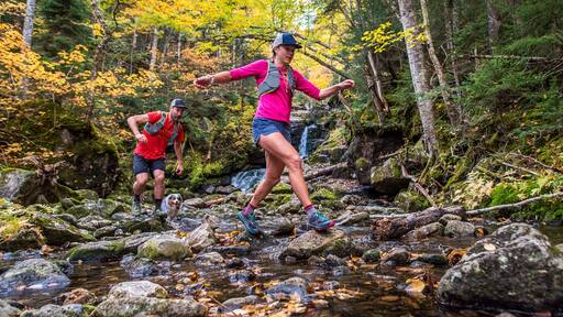 Man and woman trail runners rock-hopping in a river with fall foliage