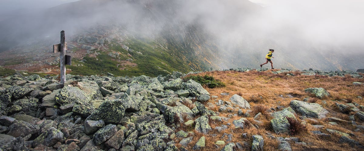 A hiker on a rocky mountain trail with a wooden signpost and mist-covered peaks in the background.